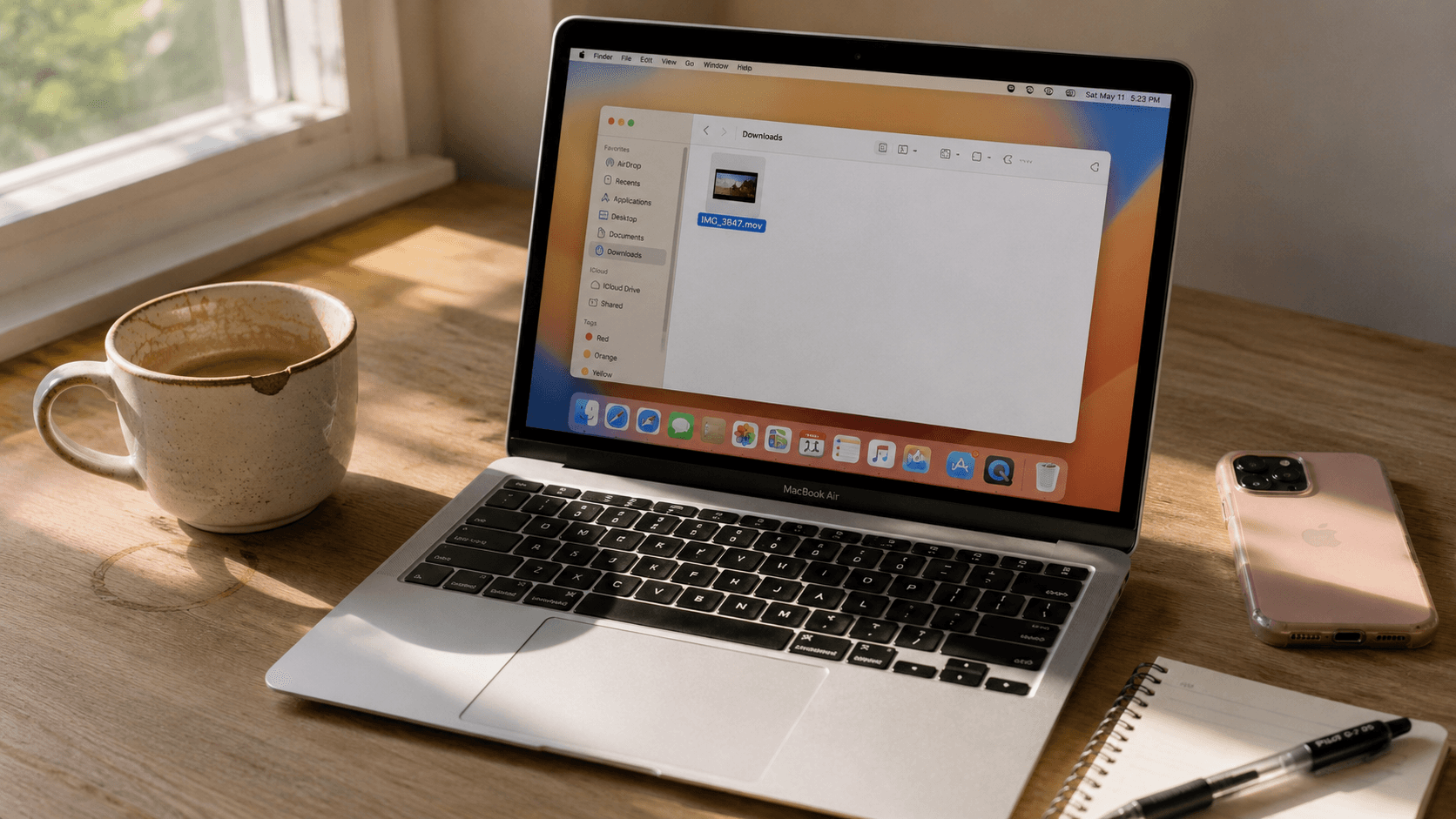 MacBook Air on a wooden desk, screen showing a Finder window with an MOV video file selected. A pink iPhone case and a coffee mug visible alongside.